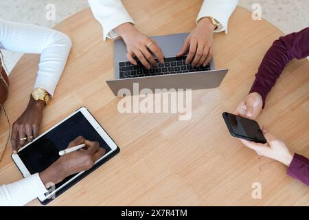 Three women of different multiethnic backgrounds are seated around a wooden table working together on digital devices, including a laptop, a tablet, a Stock Photo
