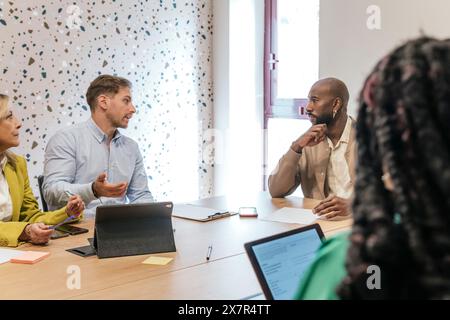 A multicultural team collaborates in a well-lit coworking office, engaged in a lively conversation with digital devices in use Stock Photo