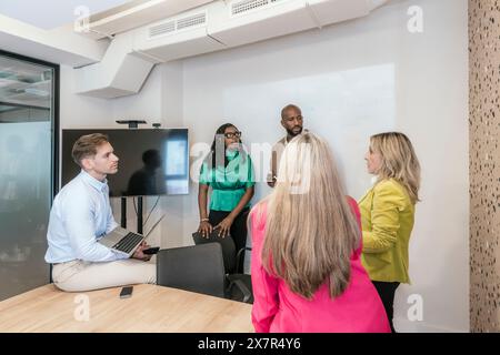 A diverse team collaborates in a modern coworking space, engaging in a productive discussion around a conference table with a television screen Stock Photo