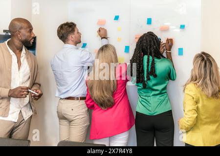 A multiethnic group of professionals collaboratively brainstorming with sticky notes on a whiteboard in a bright coworking environment Stock Photo