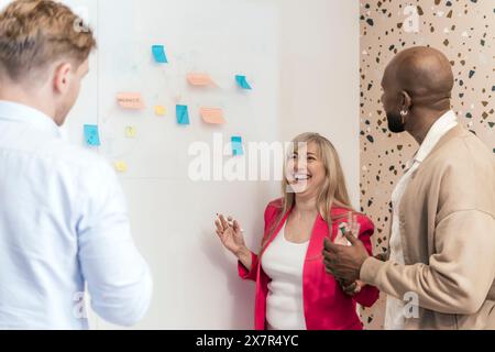 Three professionals engage in a lively brainstorming session with colorful sticky notes on a whiteboard in a coworking office setting, embodying teamw Stock Photo
