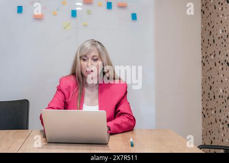A focused businesswoman in a pink blazer works diligently on her laptop at a coworking desk with sticky notes on the wall Stock Photo