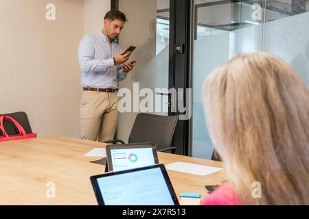 Man using smartphone stands while woman works on laptop in a bright coworking office environment, exhibiting productivity and collaboration Stock Photo