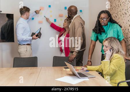 Diverse team of professionals engaging in a brainstorming session at a coworking office space, with one member taking notes and another on a phone cal Stock Photo