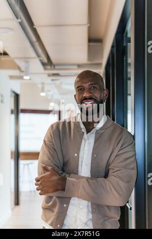 A professional and confident bald man with a beard smiles while standing in a coworking office environment with folded arms Stock Photo
