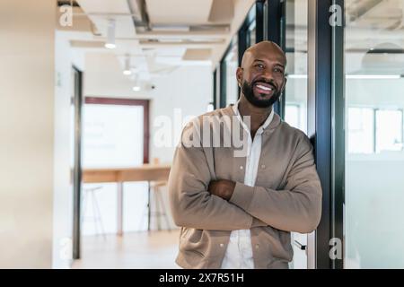 Smiling man in casual attire confidently standing with arms crossed in a well-lit coworking office, portraying a professional and approachable demeano Stock Photo