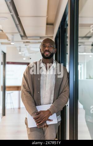 A well-dressed business professional stands contemplatively in a stylish coworking office, holding documents Stock Photo