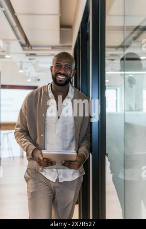 A cheerful bald man in casual business attire stands holding a digital tablet in a modern coworking office environment Stock Photo