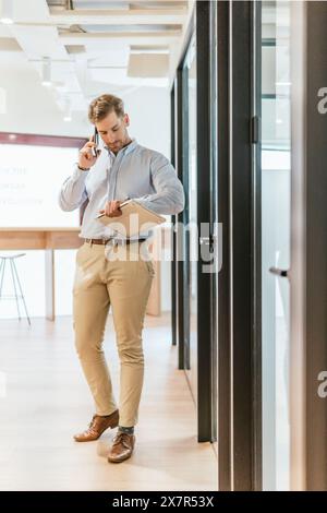 Professional male walks through a coworking office while engaged in a phone conversation and holding documents Stock Photo
