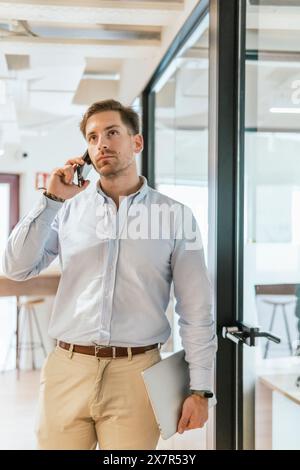 A professional in a smart casual attire is engaged in a conversation on his smartphone while holding a laptop, standing in a well-lit coworking office Stock Photo