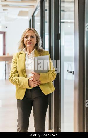 Confident mature businesswoman holding a laptop stands in a bright coworking office space, radiating professionalism and experience Stock Photo
