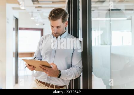 A focused professional male in a smart-casual shirt stands engrossed in reviewing his notes on a clipboard within a bright coworking office environmen Stock Photo