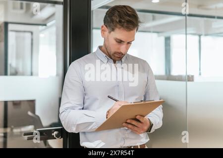 A focused young professional stands in a well-lit coworking office, diligently taking notes on a clipboard Stock Photo