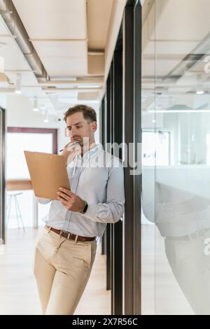 A pensive young male professional stands with a clipboard in a bright coworking office, contemplating his next business move Stock Photo