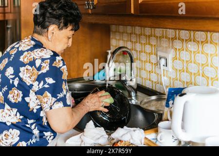 A senior woman is captured washing dishes in her homely kitchen, surrounded by cooking utensils and a vintage tiled backdrop Stock Photo