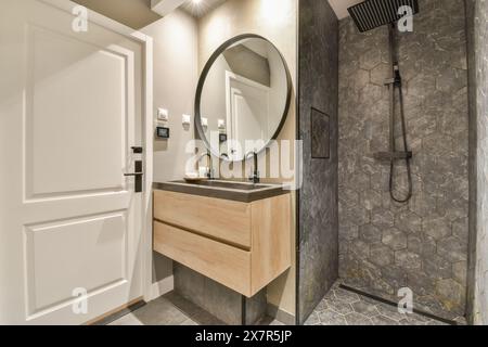 This contemporary bathroom showcases a circular mirror above a wooden vanity, set against a textured dark stone wall The space includes a minimalist s Stock Photo