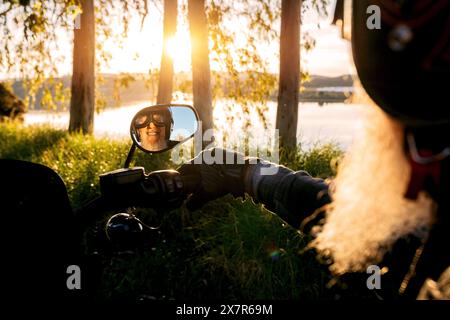A smiling bearded senior man wearing a helmet is reflected in a motorcycle's side mirror, looking at the camera with a backdrop of sunlit trees by a l Stock Photo
