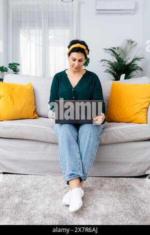 A focused young woman sits cross-legged on her sofa, deeply engaged with work on her laptop in a sunny, plant-filled living room Stock Photo