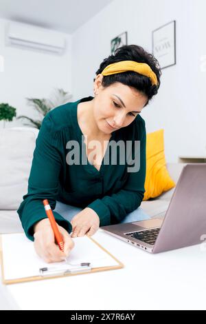 A woman in a green blouse focuses on writing notes on a clipboard while seated beside a laptop in a bright, well-decorated room Stock Photo