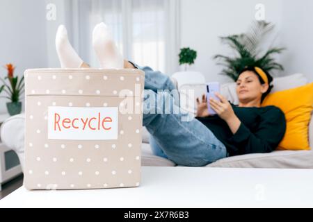 A relaxed woman lounges on her sofa using a smartphone, while a dotted box labeled Recycle rests prominently in the foreground, highlighting eco-frien Stock Photo