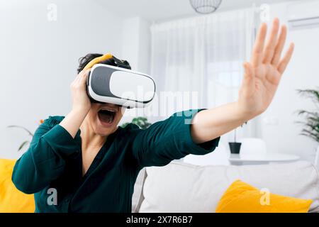A woman in a dark green shirt expresses joy while using a virtual reality headset in her bright, modern living room Stock Photo