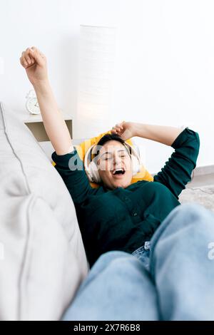 A joyful woman stretches with a big smile while wearing headphones and lying comfortably on a sofa at home Stock Photo