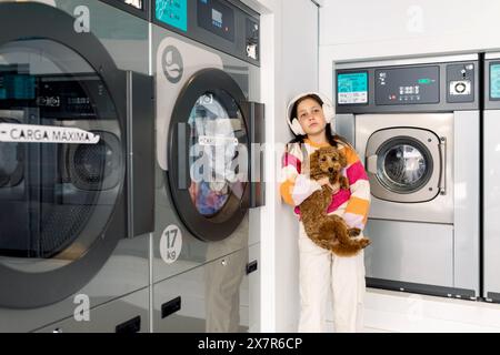 A preteen girl holding a dog stands in a laundry room with headphones on, looking away. Stock Photo