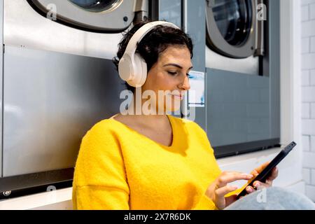 Woman with headphones listens to music and uses a mobile phone while sitting in a laundromat, surrounded by washing machines, looking down. Stock Photo