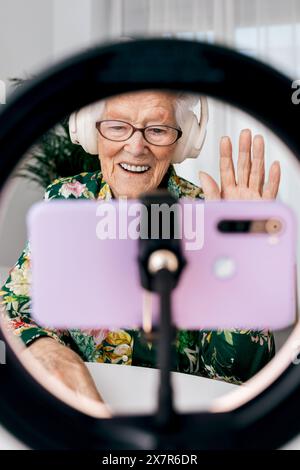A cheerful elderly woman records a podcast using a smartphone and headphones, seen through a ring light Stock Photo