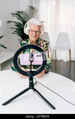 An elderly woman sits at a white table, wearing headphones and speaking into a smartphone mounted on a ring light, recording a podcast in a bright roo Stock Photo