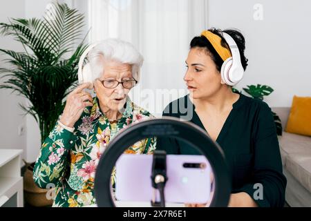 A senior woman and a younger adult female sit together recording a podcast They both wear headphones and speak into a microphone, recording with a sma Stock Photo