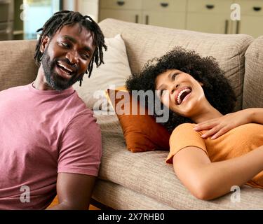 Laughing Couple At Home With Woman Lying On Sofa And Man Sitting On Floor Stock Photo