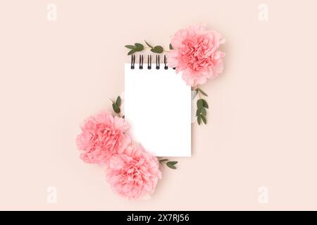Empty notepad mockup, pink carnation flowers and green eucalyptus branches on a beige background. Greeting card concept. Stock Photo
