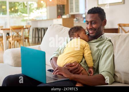 Family With Father Working On Laptop Cuddling Sleeping Baby Daughter On Sofa At Home Stock Photo