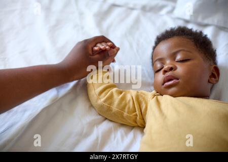 Close Up Of Loving Mother Holding Baby Daughter's Hand As She Sleeps On Bed At Home Stock Photo