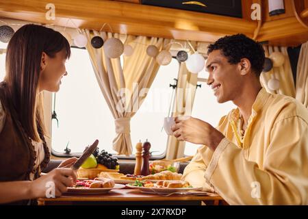 An interracial man and woman sitting at a table, enjoying a delicious meal together in a camper van on their romantic getaway. Stock Photo