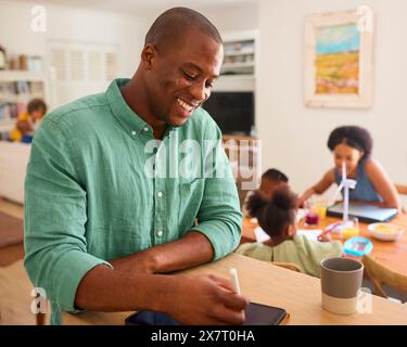 Man Working On Digital Tablet With Stylus Indoors At Home With Multi-Generation Family In Background Stock Photo