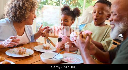 Grandparents With Grandchildren At Home Eating And Decorating Cakes With Family In Background Stock Photo