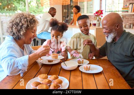 Grandparents With Grandchildren At Home Eating And Decorating Cakes With Family In Background Stock Photo