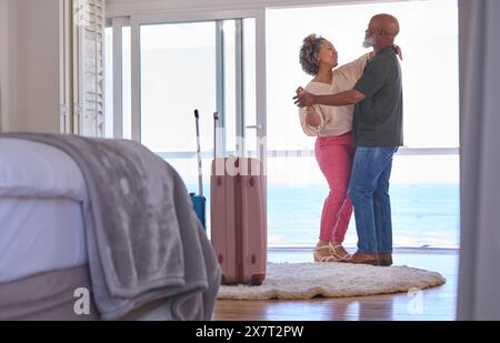 Mature Couple With Luggage Dancing In Beachfront House Overlooking Ocean For Summer Vacation Stock Photo