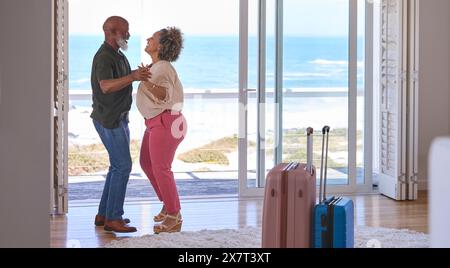 Mature Couple With Luggage Dancing In Beachfront House Overlooking Ocean For Summer Vacation Stock Photo