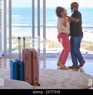 Mature Couple With Luggage Dancing In Beachfront House Overlooking Ocean For Summer Vacation Stock Photo