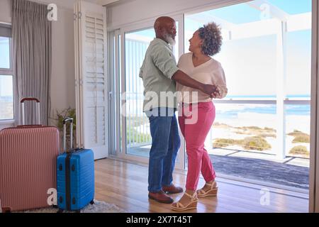 Mature Couple With Luggage Dancing In Beachfront House Overlooking Ocean For Summer Vacation Stock Photo