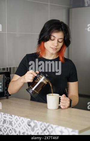 Black-haired white person serving coffe from a french press to a mug with a relaxed smiling expresson on their face. Stock Photo
