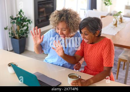 Two Mature Female Friends At Home With Laptop Making Video Call To Family Or Friends Stock Photo