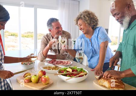 Group Of Smiling Mature Friends At Home Relaxing Preparing Food For Lunch With Wine Together Stock Photo