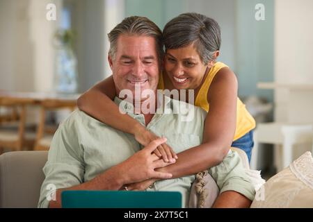 Mature Couple At Home With Woman Hugging And Surprising Man Using Laptop Sitting On Sofa Stock Photo