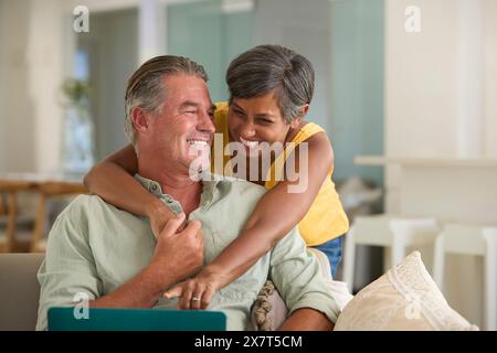 Mature Couple At Home With Woman Hugging And Surprising Man Using Laptop Sitting On Sofa Stock Photo