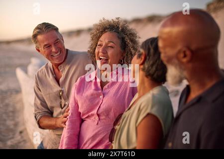 Mature Couple With Friends Sitting Outdoors On Beach Watching Sunset Together Stock Photo