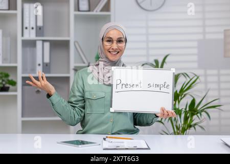 Smiling teacher in hijab holding whiteboard with 'Present Simple' written on it, explaining grammar in a modern office setting. Stock Photo